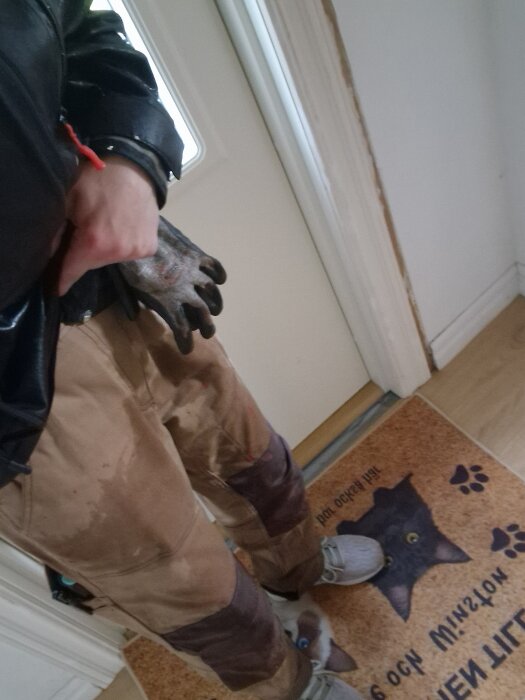 Man i arbetskläder står på en dörrmatta med kattmotiv, iförd smutsiga arbetsbyxor och handskar i handen.