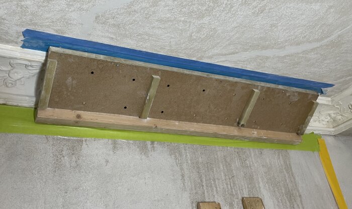 Träram med hål och stödpinnar fäst vid taket, omgiven av blå och grön tejp för renovering av gipsstukatur.
