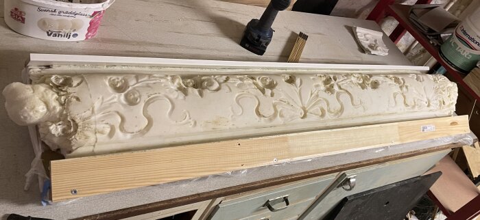 Gipsstukatur på arbetsbänk med detaljerade blommönster, omgiven av trästöd för avformning. Verktyg och material synliga i bakgrunden.
