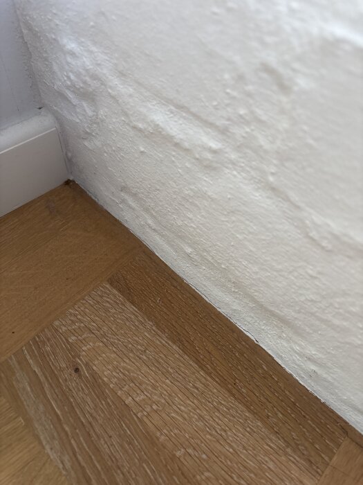 Hörn av ett rum med träparkett och vit ojämn vägg där träfärgad mjukfog applicerats i springa.