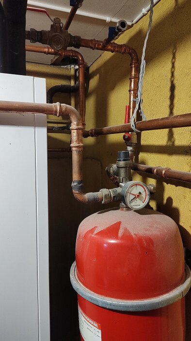 Rör och ventiler i värmesystem med rött expansionskärl och tryckmätare, blå kran tydlig, inhägnat utrymme med gul vägg.