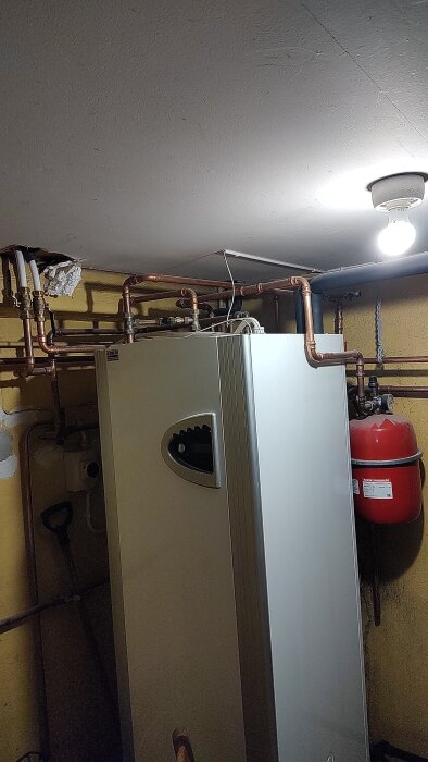 Värmesystem med flera kopparrör och ventiler i ett pannrum. En röd tryckbehållare syns till höger.