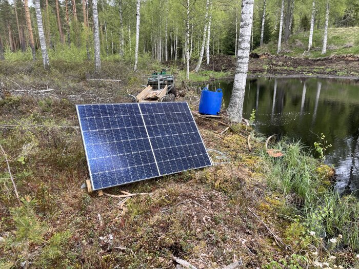 Solpanel vid damm med blå tunna kopplad för vattenrening i skogsmiljö.