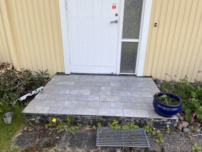 Stor, fyrkantig entrétrappa med klinkerplattor. Dörr med glasparti och sidofönster. Blomkrukor och växter runt trappan.