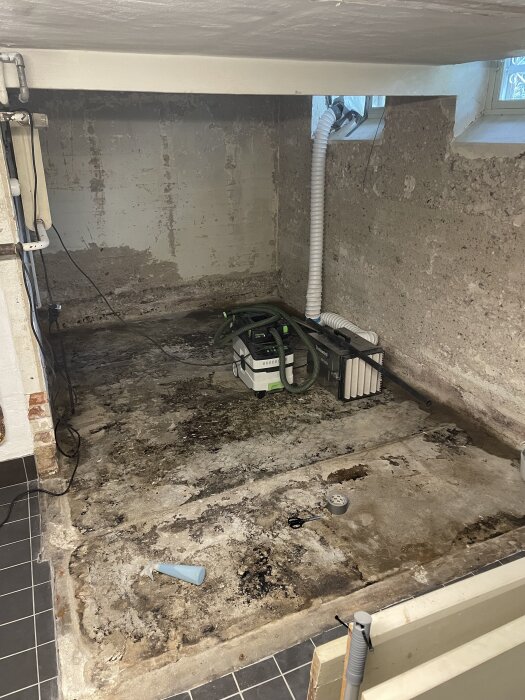 Riven golvyta i renoveringsprojekt med fuktfläckar på betongen, kvarvarande äldre material och en luftrenare med slangar i ett rum med fönster.