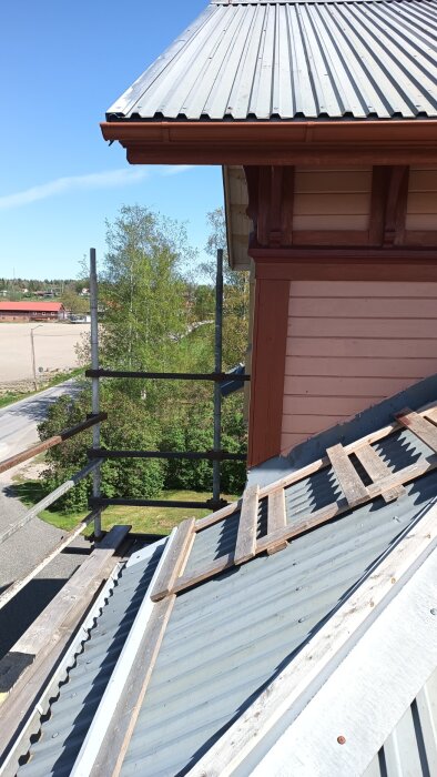 Byggställning på tak med målade fasaddetaljer, omgiven av träd och blå himmel, taget i blåsigt väder med utsikt över området.