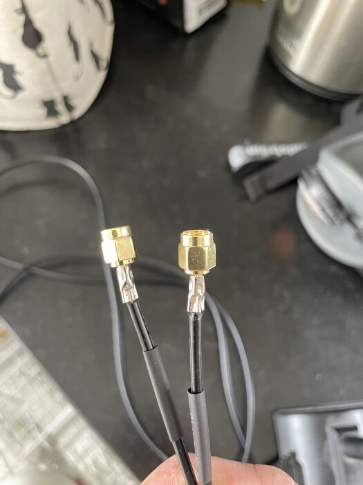 Två SMA-kontakter på svarta kablar med lödda centerstift och delvis monterade yttermantlar, hållna i en hand på en mörk yta.