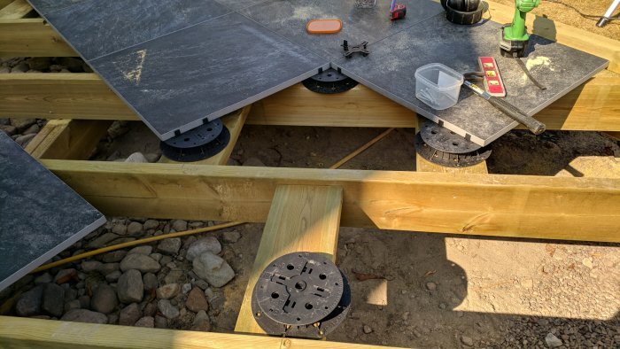 Trallbygge med pågående montering av stenplattor på plintar; verktyg och byggmaterial synliga på bilden.