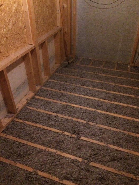 Isolering mellan träreglar i golvkonstruktion, med synliga träpaneler på väggarna i bakgrunden.