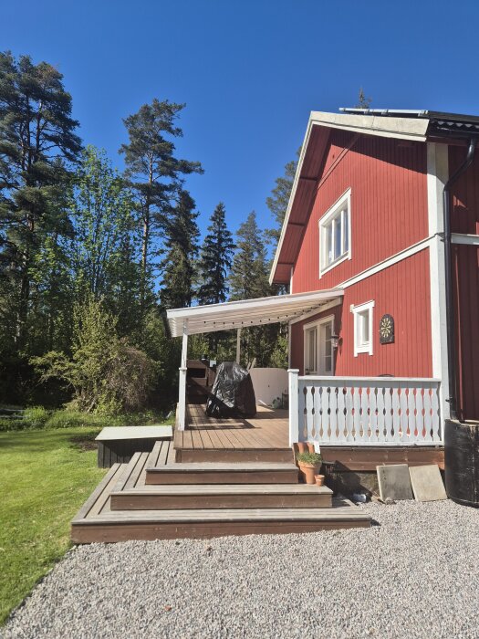 Rött trähus med vita detaljer, veranda under tak, trappsteg och grusväg. Skog i bakgrunden, blå himmel.