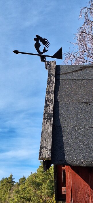 Väderflöjel med siluett av en sjöjungfru på taknock, mot en klar himmel med träd i bakgrunden.