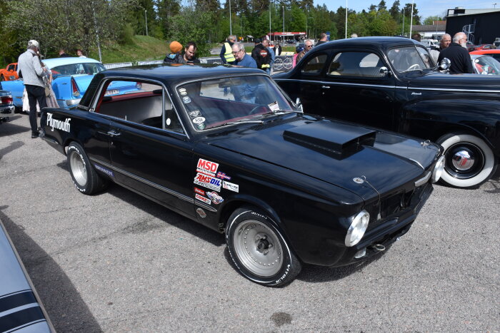 Svart Plymouth-bil på en bilträff i Båberg, Trollhättan, omgiven av andra bilar och besökare.