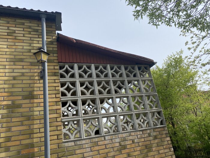 Tegelvägg med trapetsformat mönster och lampa, behöver renovering. Renoveringsarbeten planeras för vindskivor och entrétak.