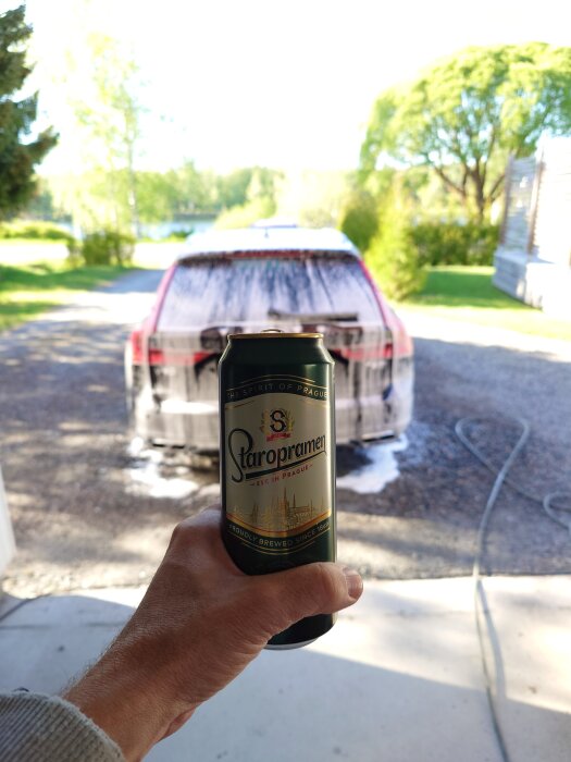 En person håller en burk Staropramen-öl i förgrunden med en bil under biltvätt i bakgrunden på en solig dag.
