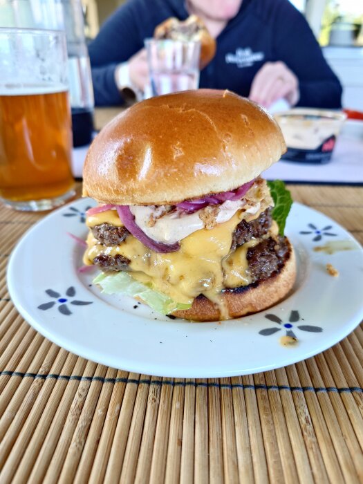 Dubbel ostburgare med picklad rödlök, rostad lök, gul senap, Max Sourcreme Pepper, sallad och tomat på en tallrik, med öl i bakgrunden.