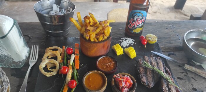 Tallrik med biff, pommes frites, lökringar, grillade grönsaker, majs, såser och en flaska Safari öl på Coco Beach, Dar es Salaam.