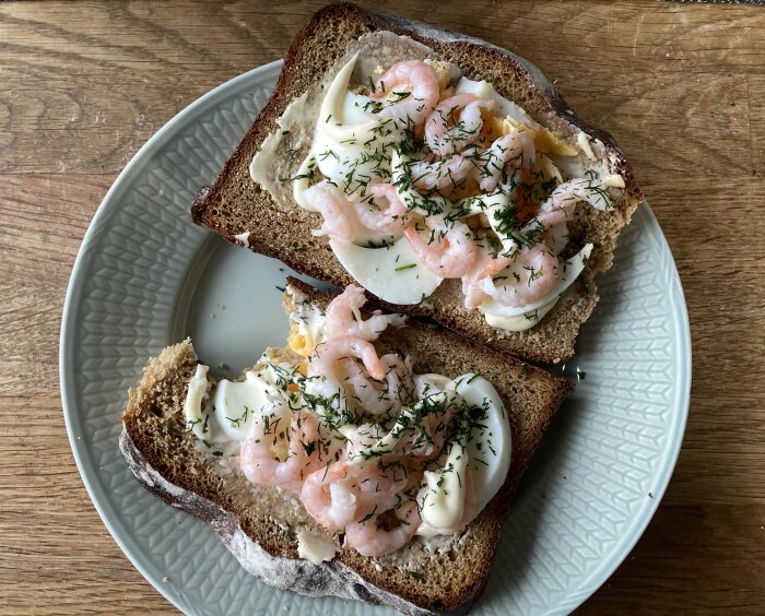 Två räkmackor med ägg, majonnäs och dill på mörkt bröd på en tallrik.