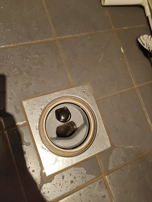 Golvbrunn i badrum med fukt på klinkers, exponering av rött textilliknande tätskikt syns delvis ovanför golvbrunnen.