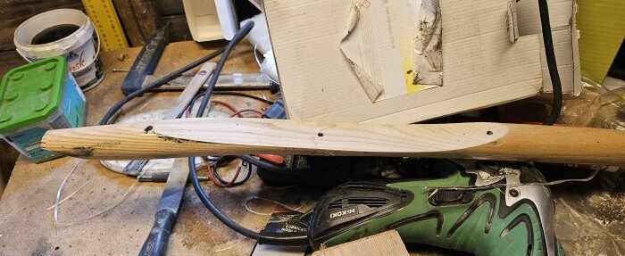 Ett trasigt skaft av en spade i trä ligger på en arbetsbänk, omgivet av verktyg och en kartong.