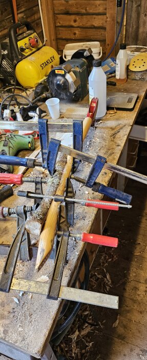 Spadhandtag lagas och förstärks på en arbetsbänk med klämmor och lim, omgiven av verktyg som slipmaskin och skruvmejsel.