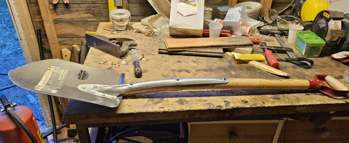 En reparerad Fiskars spade med förstärkt skaft på en arbetsbänk omgiven av verktyg och träbitar i en verkstad.