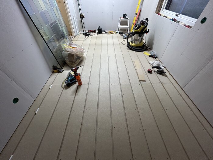 Renoverad vägg och golv av gips i ett rum med verktyg och material för golvvärmeinstallation.