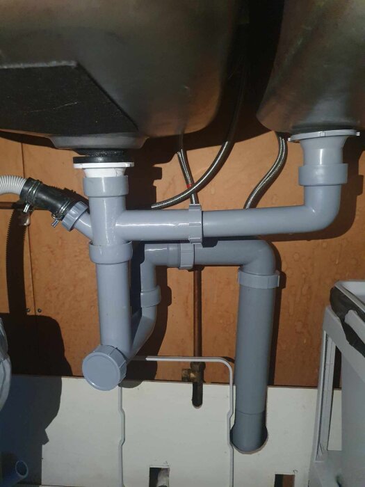 Diskbänk med grå avloppsrörsystem, inklusive sprucken vit komponent vid anslutningen, vilket orsakar läckage under diskhon.