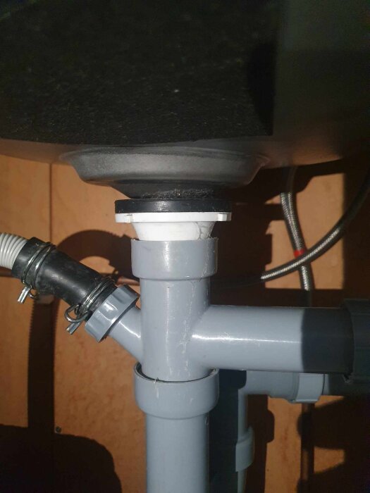 Sprucken vit del under diskhon där avloppet ansluter, orsakar läckage; visas med gråa rörledningar och slangklämmor.