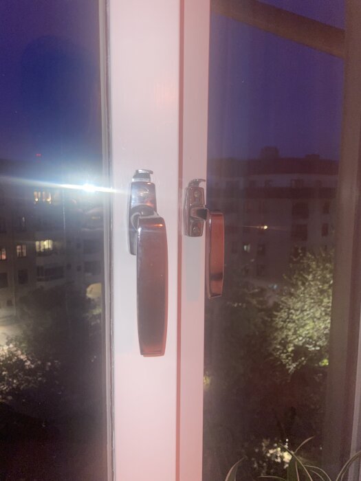 Två fönsterhandtag av metall med låspigg syns i skymningens ljus mot en bakgrund av byggnader och träd.
