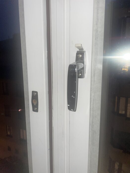 Fönsterhandtag med låspigg på vitt fönster i kvällsbelysning, del av diskussion om att byta till enhetliga mässingsfärgade handtag utan låspigg.