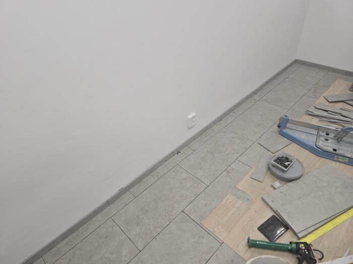 Grå keramiska lister mot en vit vägg med verktyg och material på golvet, redo för plattsättning och fogning i ett maskinrum.