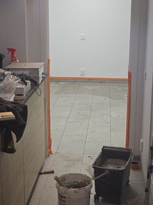 Rum under renovering med kaklade golv, upptagna lister och verktyg på golvet. En hink och andra renoveringsmaterial syns i förgrunden.