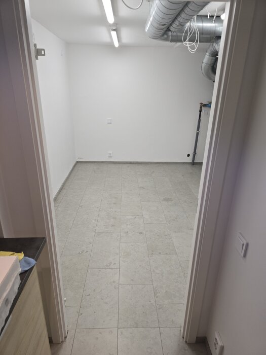 Ett ljust rum med kakelgolv, vit vägg och synliga ventilationsrör i taket, upplyst av två långa ljusarmaturer.