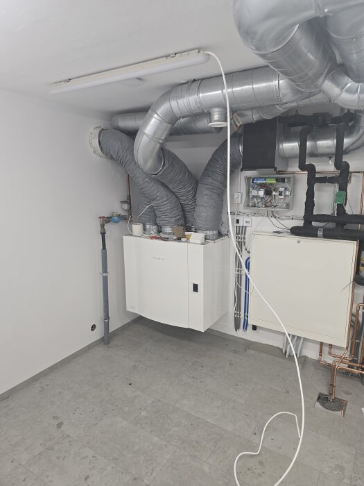 Ventilationssystem och rör i ett maskinrum, med ljusgrå klinkergolv och synliga kablar på väggen. Takbelysning ger jämnt ljus.