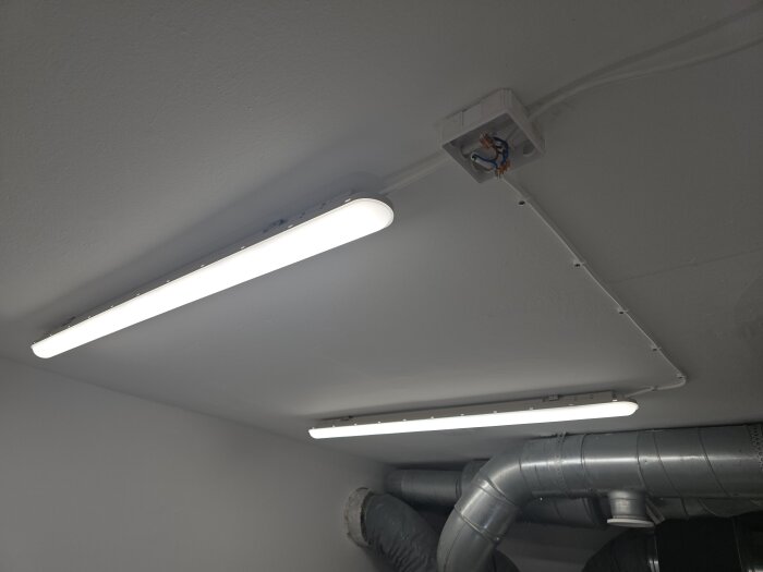 Tak med två lysrör och synliga ventilationsrör i ett maskinrum, belysningen ger jämnt ljus i rummet.