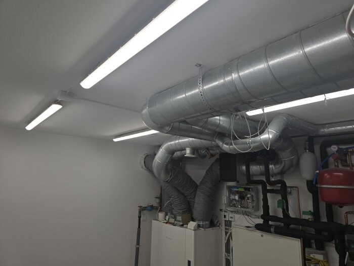 Källarrum med fyra lysrör i taket, stora ventilationsrör och teknisk utrustning, kraftigt upplyst trots att rummet saknar fönster.