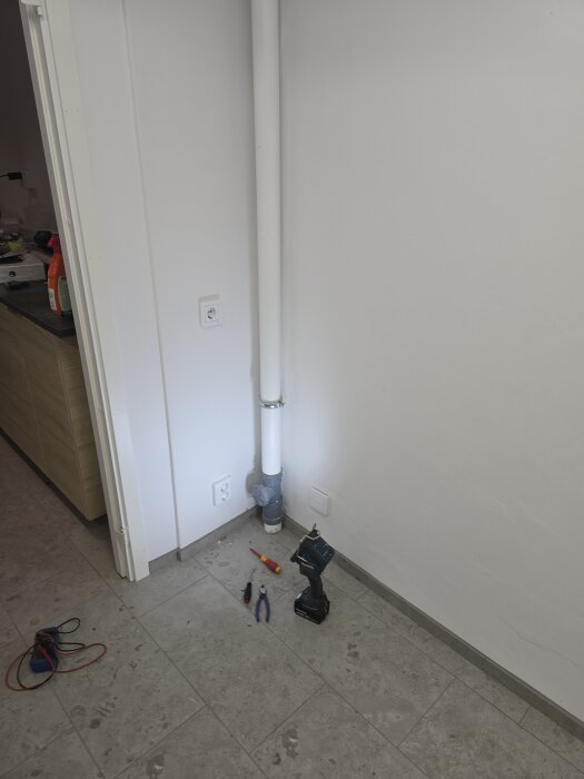 Verktyg och en borrmaskin på golvet vid ett vitt rör som går längs väggen. Elektriska dosor syns intill röranslutningen.
