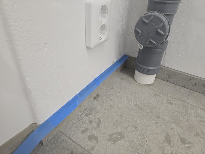 Vägguttag installerat nära en grå rörledning, med blå tejp längs väggen på ett grått tegelgolv.
