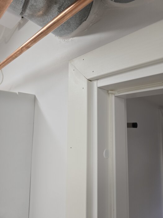 Dörrkarm i ett rum med vitmålad dörrpost, synlig kopparledning och isolerad ventilationsrördetalj i taket.