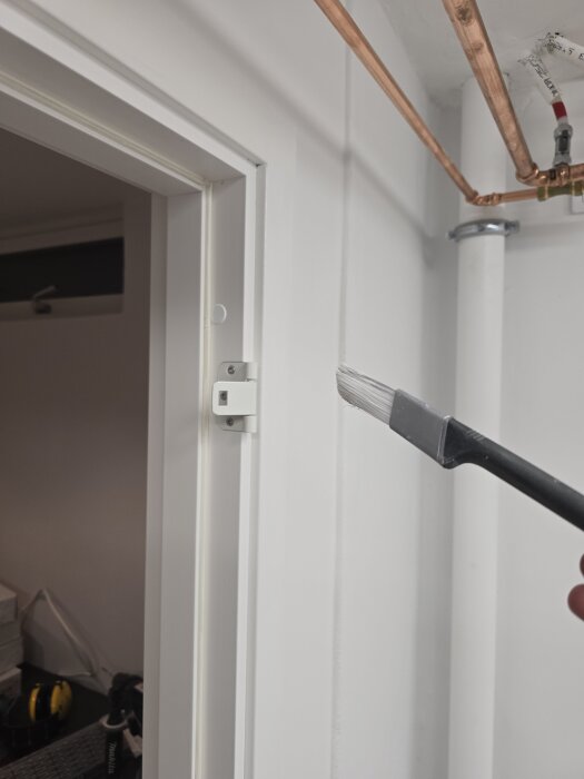 Pensel applicerar vit färg på dörrfoder, omgivande synliga kopparrör och eldetaljer.