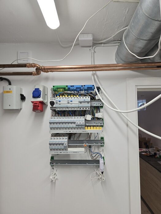 Elcentral med flera säkringar och kablar i en vitmålad vägg, kopparrör och ventilationsrör synliga ovanför.