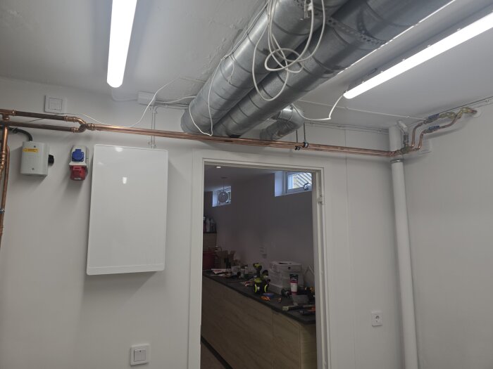 Elinstallation i maskinrum med rörledningar, kablar, elcentral och kopplingsuttag på väggen. Rummet har vita väggar och trälist.