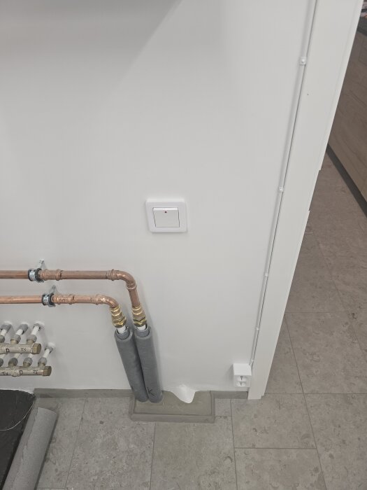 Vägg med kopparrör, ventiler och isolerade vattenrör, samt ett uttag och en strömbrytare på en ljus vägg i ett rum med ljusgrått klinkergolv.