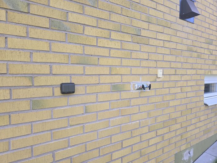 Gul tegelvägg med nyinstallerat utomhusuttag, vattenutkastare och yttertempgivare nära ett fönster.