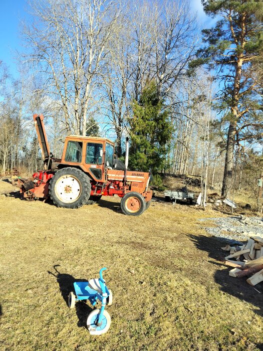 Röd Belarus 800 traktor från 1983 på gräsmatta med träd i bakgrunden, en blå trehjuling syns i förgrunden.
