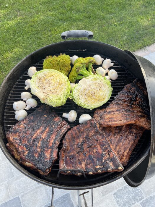 Grillad mat på kolgrill: revben, halverad broccoli och kål samt champinjoner. Bakgrund av grön gräsmatta och stenplattor.