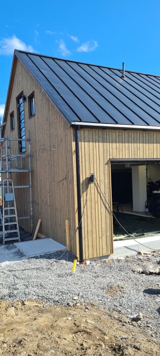 Byggnad med träfasad behandlad med lasyr Gråtrut, stege och byggställning synliga mot blå himmel.