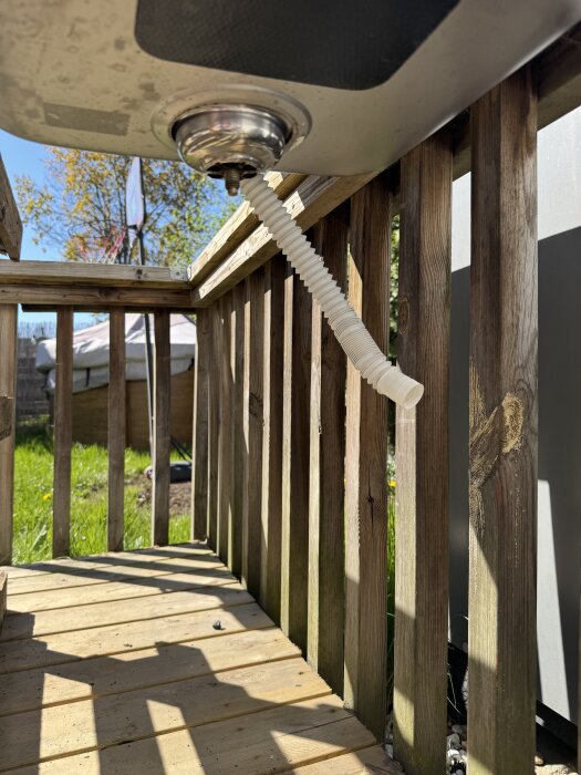 Utekök underifrån med synlig avloppsslang och diskho, monterad på en altan av trä med staket i dagsljus.