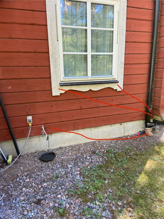 Röd husfasad med ett fönster och två röda pilar som pekar mot marken. Området är av grus och gräs. Plan för dränering av vatten.