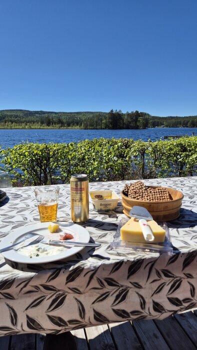 Lunchbord vid sjö i solsken, med sill, potatis, gräddfil, knäckebröd med ost, Bregott, och en öl på en duk med lövmönster. Utsikt över vatten och skog.